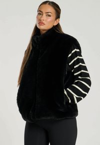 Svart fuskpälsväst med hög krage och randiga långärmar i svart och vitt. Slät textur, avslappnad passform och ingen synlig hårdvara.