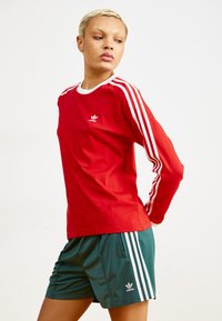 Rød langermet skjorte med hvit krage og tre hvite striper på ermene, sammen med grønne shorts med tre hvite striper og logo.