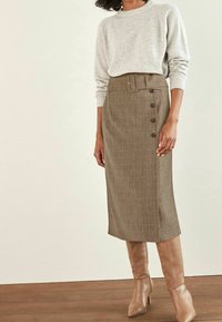 Femme portant un pull en tricot beige rentré dans une jupe midi marron ceinturée avec des boutons sur le côté et des bottes à talons hautes couleur fauve.