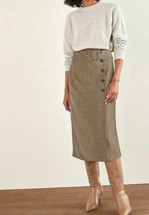 Žena v béžovém pleteném svetru zastrčeném do hnědé midi sukně s páskem a bočními knoflíky, doplněná světlými kozačkami na vysokém podpatku.