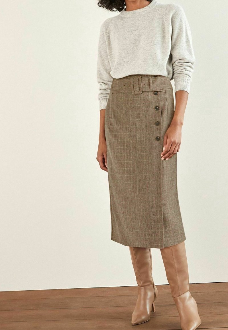 Femme portant un pull en tricot beige rentré dans une jupe midi marron ceinturée avec des boutons sur le côté et des bottes à talons hautes couleur fauve.