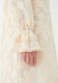 Vestido de flores en crema con tejido texturizado, puños con volante que presentan un borde en forma de concha y una sobrecapa semi-translúcida.