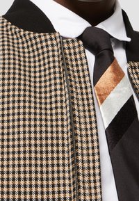 Chemise à col blanc avec une cravate rayée noire, marron et blanche portée sous une veste zippée marron et noire à carreaux.