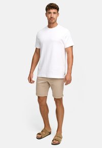 T-shirt en coton blanc, shorts en tricot beige, et sandales à enfiler marron. Les shorts sont retroussés au niveau de l'ourlet, mettant en valeur une coupe décontractée et confortable.