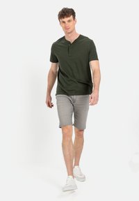 Ciemnozielona koszulka henley z krótkim rękawem i trzema guzikami, w połączeniu z jasnoszarymi szortami dżinsowymi i białymi trampkami. Gładki materiał, luźny fason.