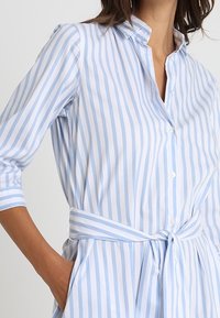 Abito a righe in azzurro chiaro e bianco, con colletto a camicia, maniche a tre quarti e una cintura legata in vita.