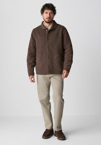 Braune Wollmischungjacke mit Kragen, Knopfverschluss, Seitentaschen, kombiniert mit hellbeigen Hosen und braunen Wildlederschuhen.