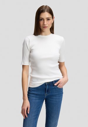 Jeune femme aux cheveux bruns et raides portant un haut blanc côtelé à manches courtes et un jean bleu, debout devant un fond gris uni.