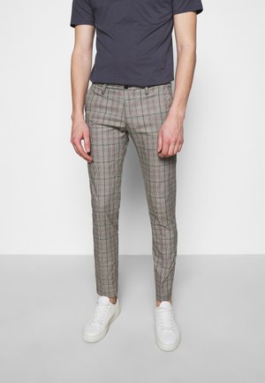 Hombre con pantalones ajustados de cuadros grises, camiseta azul marino y zapatillas blancas, de pie contra un fondo claro y liso.