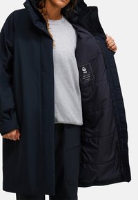 Parka impermeabile di colore blu scuro con cappuccio, dotata di chiusura con zip frontale, fodera trapuntata e un'etichetta all'interno. Il modello indossa una felpa grigia sotto.