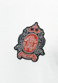 Κεντητό κόκκινο και μαύρο περίτεχνο έμβλημα με λευκές λεπτομέρειες και κείμενο «cavalli UOMO» σε φόντο λευκού υφάσματος.