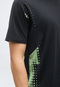 Μαύρο κοντομάνικο πουκάμισο με πράσινο pixel μοτίβο στην πλαϊνή πινελιά, φορεμένο, με εμφανές μπράτσο και ώμο ατόμου.