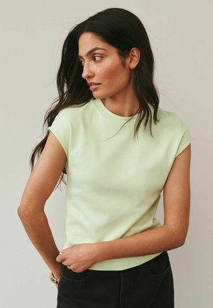 Kvinde med langt mørkt hår iført en lysegrøn kortærmet skjorte, der ser mod venstre, mens hun med den ene hånd justerer sine sorte bukser.