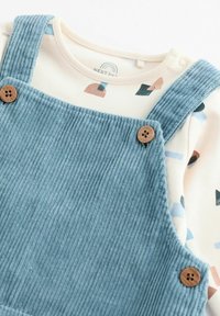 Blue corduroy overalls with button accents over a cream long-sleeve shirt featuring abstract geometric patterns in muted colors.
