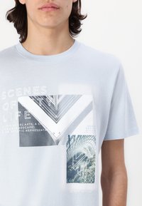 T-shirt en coton bleu clair avec des impressions graphiques de paysages architecturaux et du texte. Col rond, manches courtes et coupe décontractée.