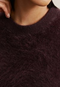 Chlupatý, tmavě burgundský svetr s kulatým výstřihem. Měkká textura s viditelnými vlákny; žebrovaný límec a mírně volný střih.