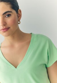 Heleroheline V-kaelusega T-särk, valmistatud pehmest kangast, lühikeste varrukatega ja lõdva lõikega. Mudelil on nähtav kuldne rõngaskõrvarõngas.