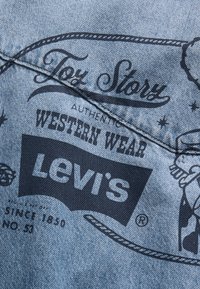 Lys blå denim med svart trykt tekst som viser "Levi's" og western-inspirerte grafikker. Glatt tekstur, uformelt design og merkbar søm.