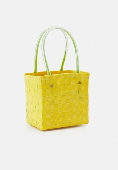 Saco tote amarelo trançado com padrão quadrado, apresentando alças verdes e tachas metálicas prateadas nas laterais. Textura suave e formato estruturado.