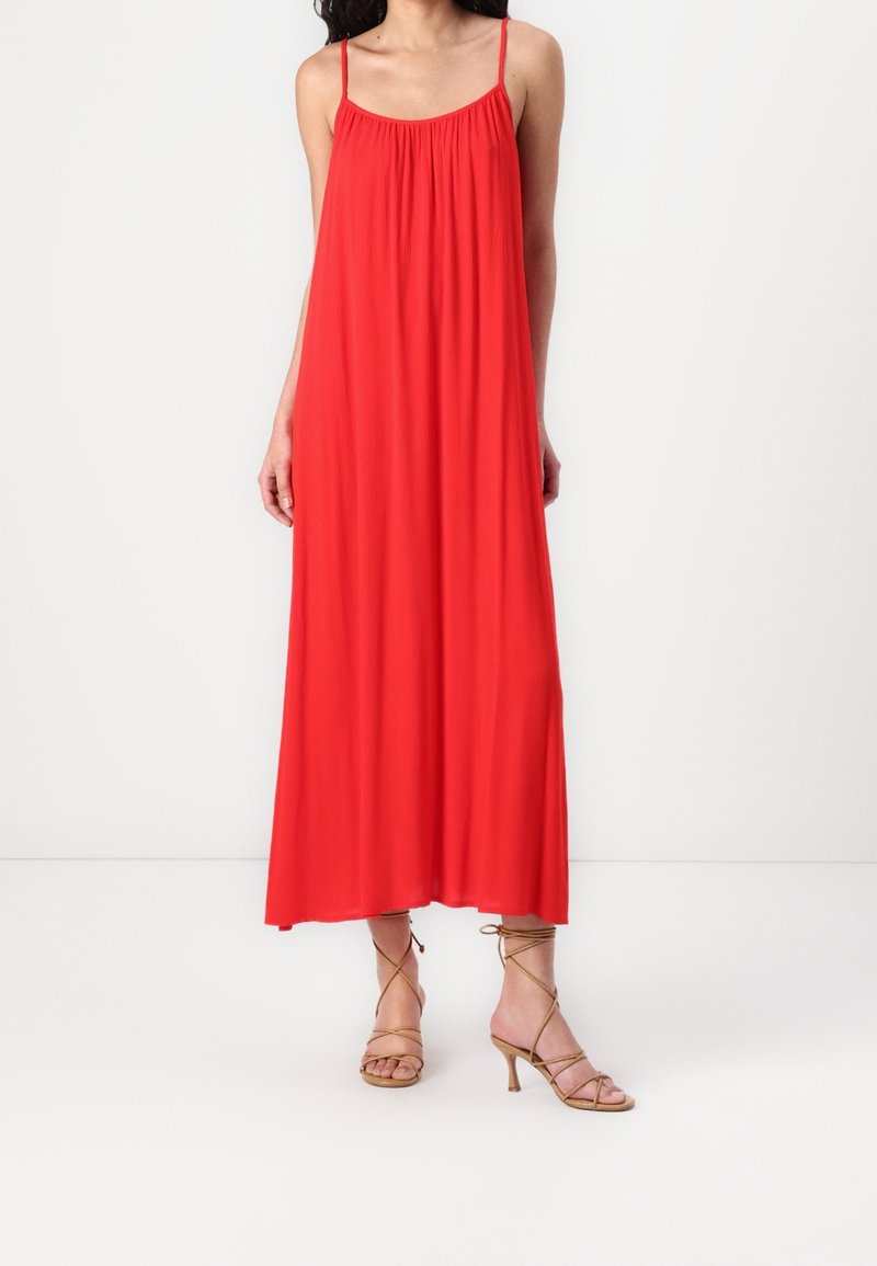 Robe longue rouge avec un col froncé et des bretelles fines, en tissu léger. Associée à des sandales à talons beiges à brides.