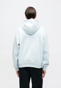 Sweat à capuche ample bleu clair avec capuche à cordon, poignets et ourlet côtelés. Le dos présente une texture lisse et mate sans logo visible.