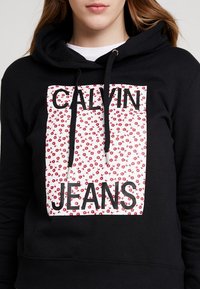 Sudadera negra con gráfico frontal que presenta flores rojas y el texto "CALVIN JEANS" en letras negras y en negrita. Material de mezcla de algodón.