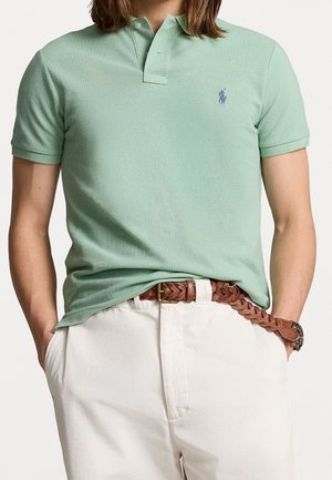 Persona que lleva una camisa polo verde claro de manga corta con un pequeño logo y pantalones blancos con un cinturón trenzado marrón, manos en los bolsillos.