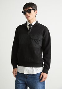 Felpa nera con mezza zip, dotata di tasca frontale e pannello testurizzato, abbinata a jeans in denim scuro.