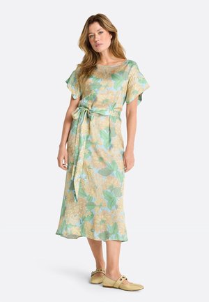 Žena v pastelovo zelenej midišate s kvetinovým vzorom, krátkymi rukávmi a opaskom na viazanie v páse, v kombinácii s béžovými plochými topánkami so cvočkami.