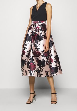 Vrouw draagt een mouwloos zwart topje en een hooggetailleerde roze satijnen rok met een groot donker bloemmotief en zwarte enkellaarzen met riempje.