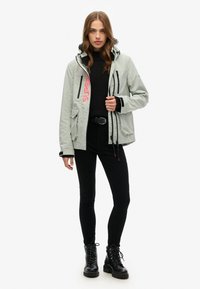 Lichtgrijze waterdichte jas met capuchon, meerdere zakken en rode "SUPER" opdruk. Draag een zwarte sweater en zwarte broek, afgewerkt met veterschoenen.