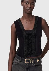 Czarny zamszowy crop top z głębokim dekoltem w serek i sznurowanym przodem. Posiada dekoracyjną czarną lamówkę i jest zestawiony z czarnym paskiem oraz szarymi dżinsami.