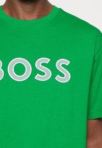 Zelené bavlněné tričko s velkým bílým perforovaným nápisem "BOSS" na hrudi. Kulatý výstřih a krátké rukávy. Měkká textura.