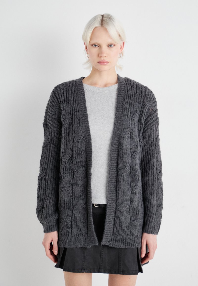 Dunkelgrau oversized Cardigan mit Zopfmuster, langen Ärmeln, offenem Front und strukturiertem Stoff, über einem hellgrauen Oberteil getragen.