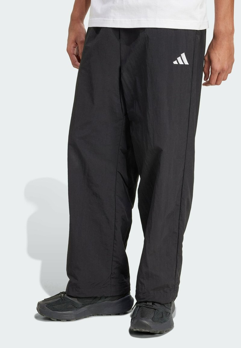 Calças pretas de nylon com um corte largo, apresentando o logótipo da Adidas em branco na perna esquerda. Textura suave e tornozelos ajustados.