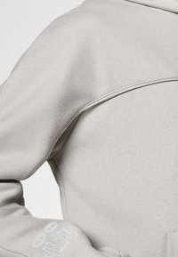Sweatshirt à capuche gris clair avec une texture lisse, présentant un détail de couture sur les épaules et un motif imprimé sur la manche gauche.