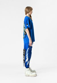 Camiseta azul de gran tamaño con gráfico en verde y negro; pantalones de chándal azules con diseño de alas blancas; botas gruesas blancas; etiqueta en los pantalones.