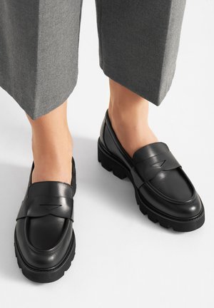 Schwarze Leder-Loafer mit abgerundeter Spitze, ausgestattet mit einem auffälligen Riemen und einer Gummisohlenprofilsohle, neben grauen, kurz geschnittenen Hosen platziert.