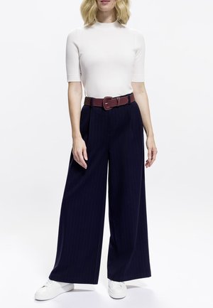 White fitted top, wide navy pinstriped trousers, brown belt with a rectangular buckle, and white sneakers. Plain background.