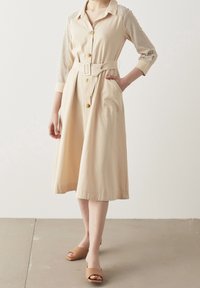 Abito beige con bottoni, vita con cintura, maniche lunghe a righe e gonna fino al ginocchio. Tessuto morbido con una leggera lucentezza.