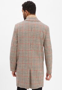 Mann, der sich abkehrt und einen beigen karierten langen Mantel mit roten und schwarzen Linien, schwarze Hose und einen beigen Rollkragenpullover trägt.