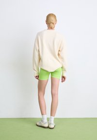 Crèmekleurige oversized trui, felgroene fietsbroek, witte sokken en multikleurige sneakers. Effen witte achtergrond en groene vloer.