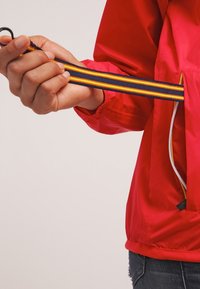 Czerwona, lekka kurtka z zamkiem błyskawicznym, z kieszenią na suwak, przedstawiająca rękę trzymającą ciemnoniebieski pasek w pomarańczowe i żółte paski. Gładka faktura.