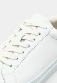White leather sneaker with flat white laces, smooth textured surface, minimal design, and subtle stitching detail along the edge.