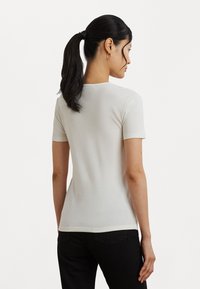 Kortärmad, ljusbeige figurnära t-shirt med rund halsringning, slät textur och rak fåll, bärs tillsammans med mörka jeans.