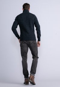Maglione lavorato a maglia blu scuro con colletto alto, indossato con jeans grigi e stivaletti in pelle marrone, che mostra una texture a coste e un design attillato.