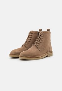 Botas de tobillo de ante marrón con diseño de cordones, suela de goma y costuras en contraste. Incluyen una pestaña en el talón para ponerlas y quitarlas fácilmente.