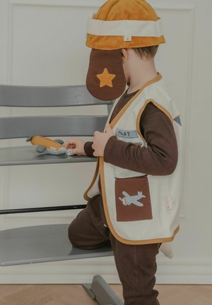 Lapsi, joka on pukeutunut lentäjän asuun, lentäjän hattuun ja liiviin, leikkii pienellä lelu lentokoneella harmaalla tuolilla sisätiloissa.