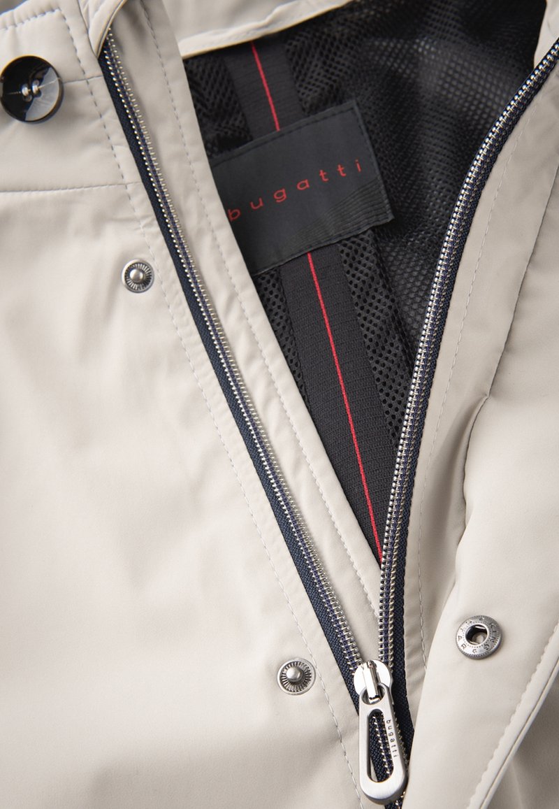 Veste beige clair avec fermeture éclair, dotée d'une doublure en maille noire et d'accents rouges sur l'étiquette. Quincaillerie en argent sur la fermeture éclair et les boutons-pression visible.