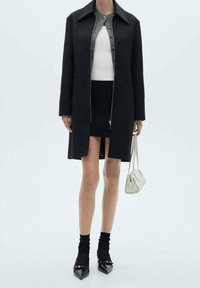 Sort lang frakke med krave og frontlynlås; parret med en hvid tætsiddende top, sort miniskørt, sorte sokker og spidse sko.